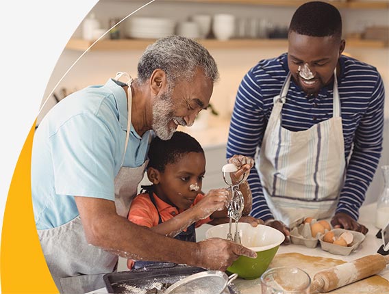 A young boy and his father and grandfather laugh together as they messily mix cake batter at the kitchen counter