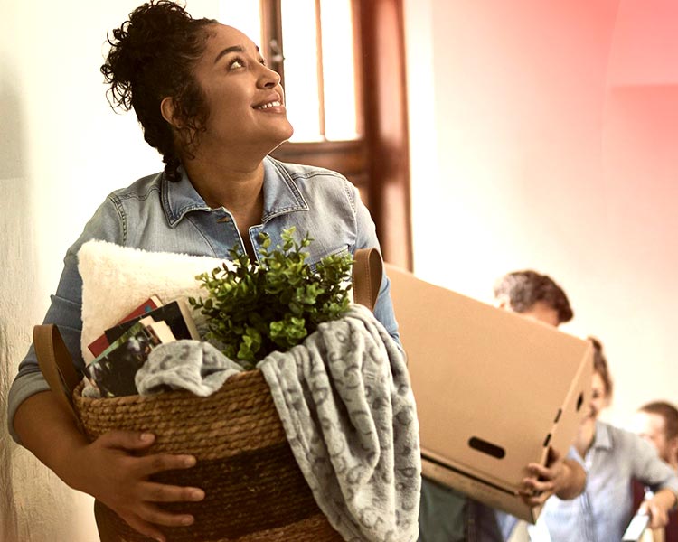A woman moving into her new home looks happy as she carries a laundry basket full of household items up the stairs